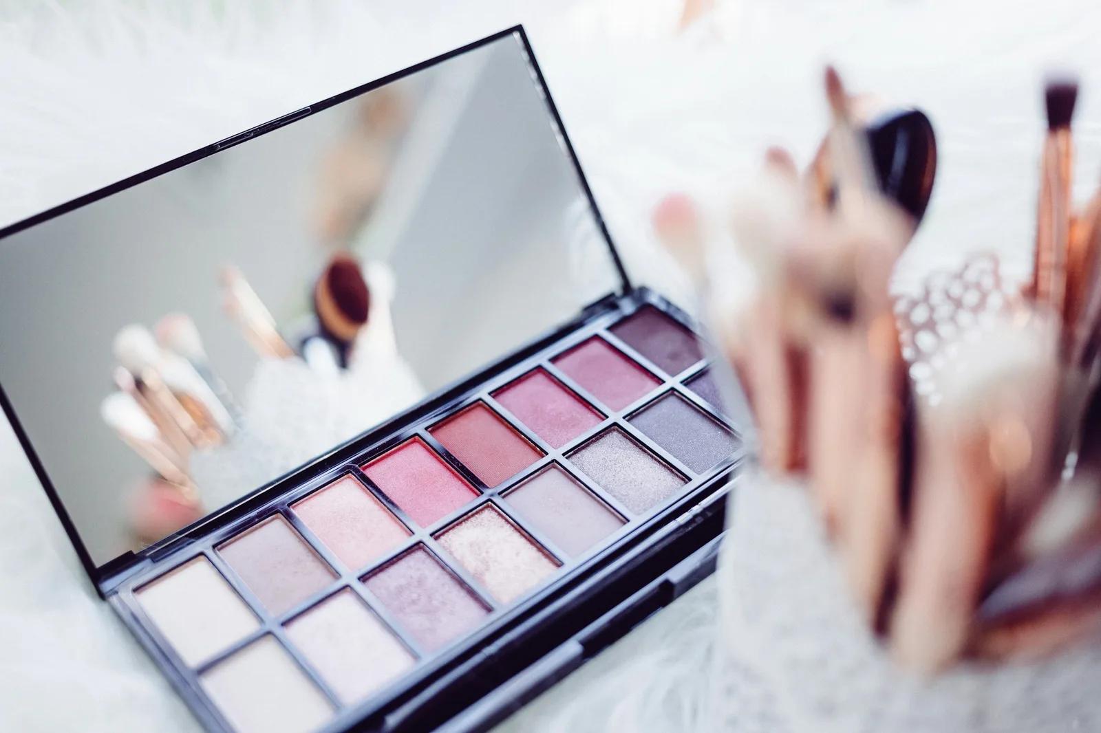 Una paleta de sombras abierta en tonos rosa suave junto a un vaso con brochas de maquillaje
