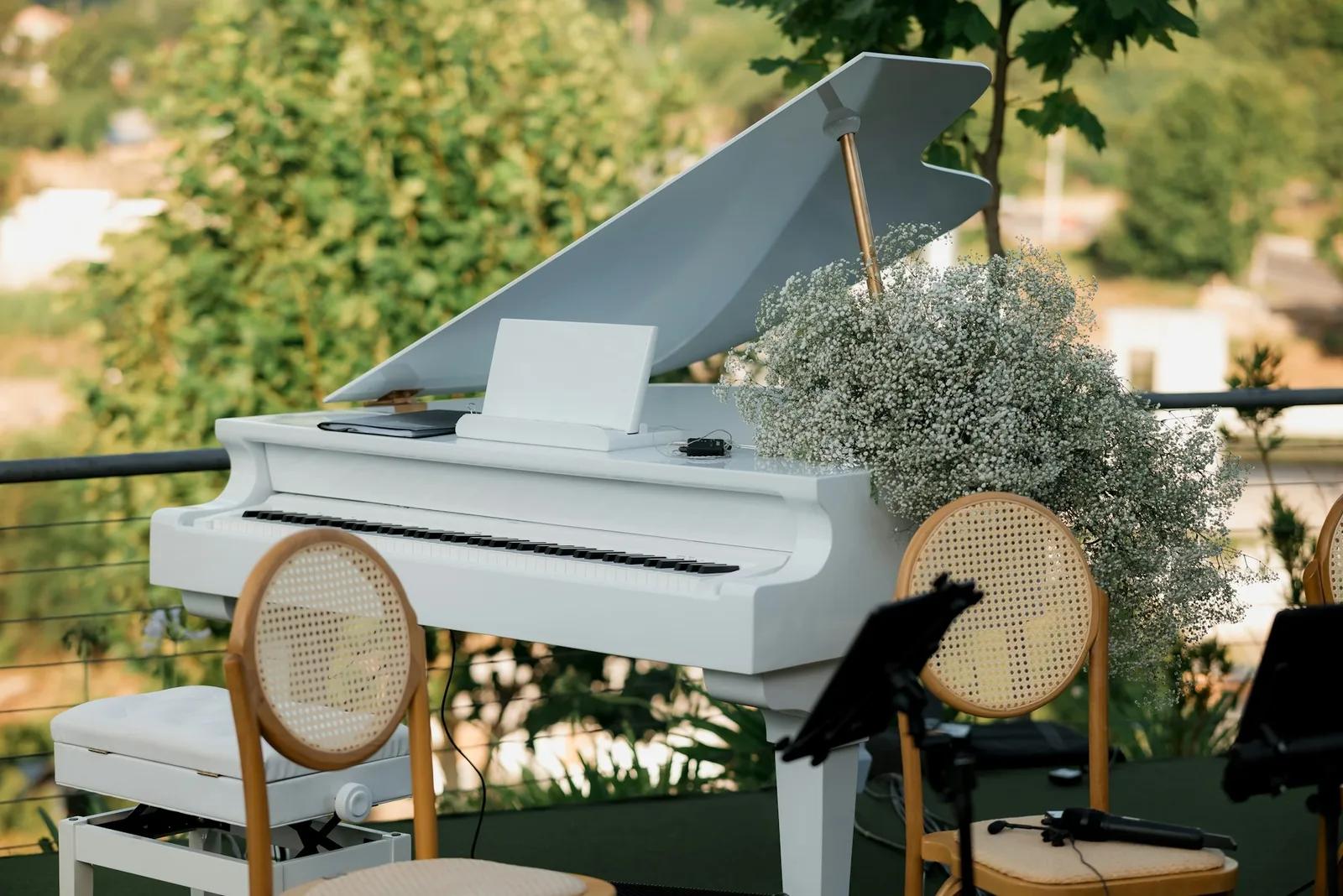 Un piano de cola blanco montado al aire libre para una ceremonia, con paniculata, sillas de ratán, atriles y un micrófono