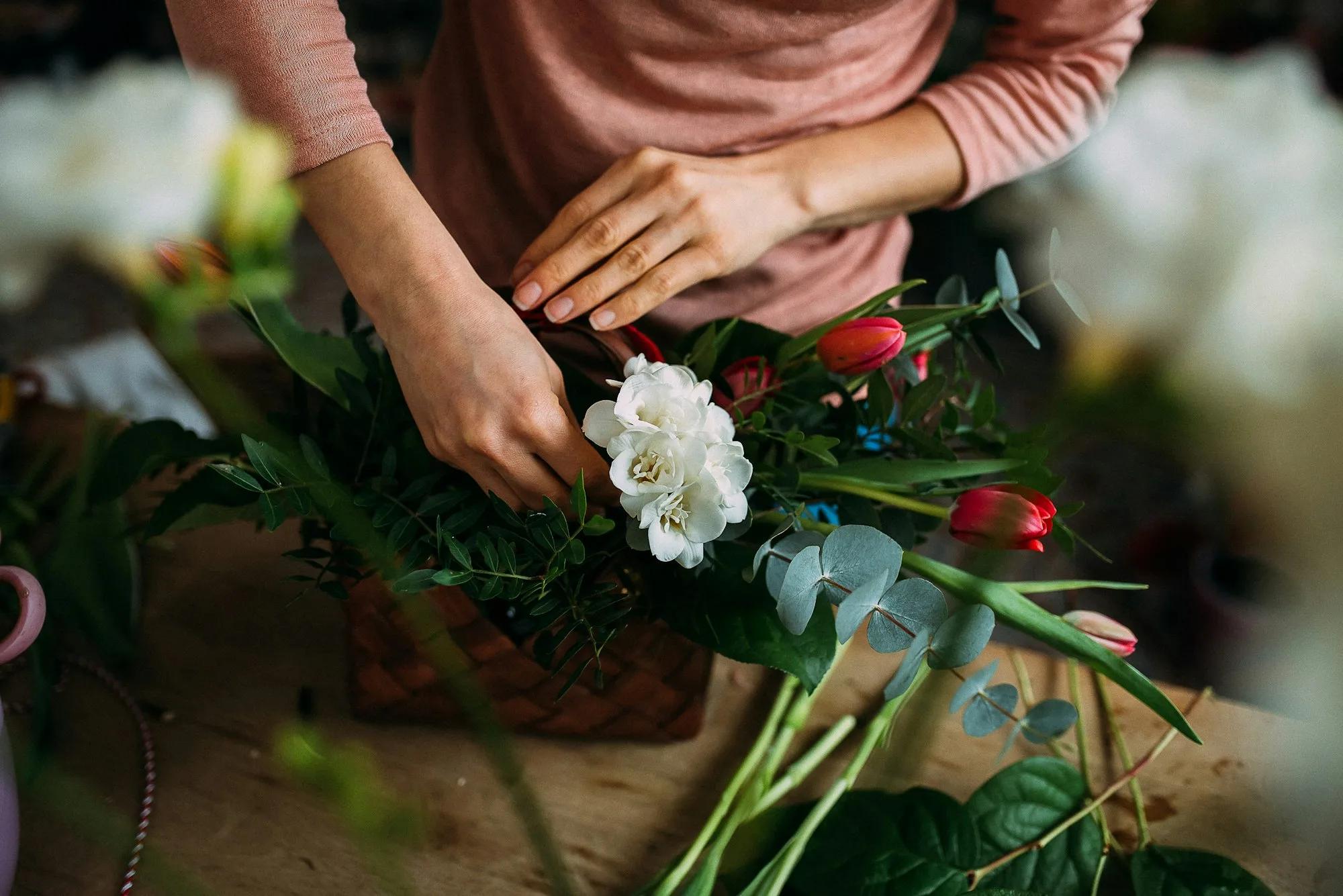 Las manos de una florista componen fresias blancas, tulipanes rojos y eucalipto en una cesta sobre un banco de trabajo de madera.