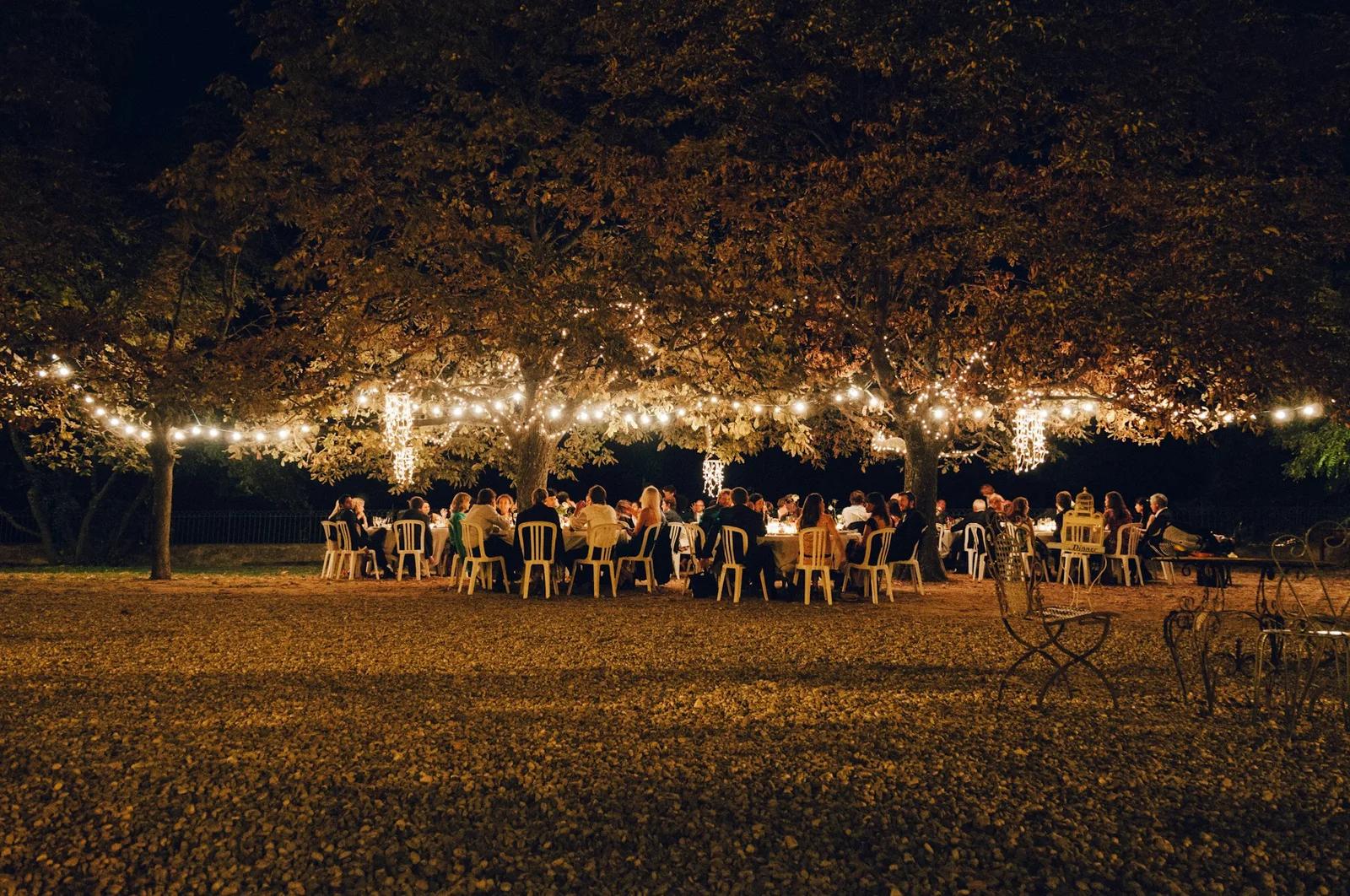 Invitados de boda sentados al anochecer en largas mesas bajo los árboles, con guirnaldas de luces