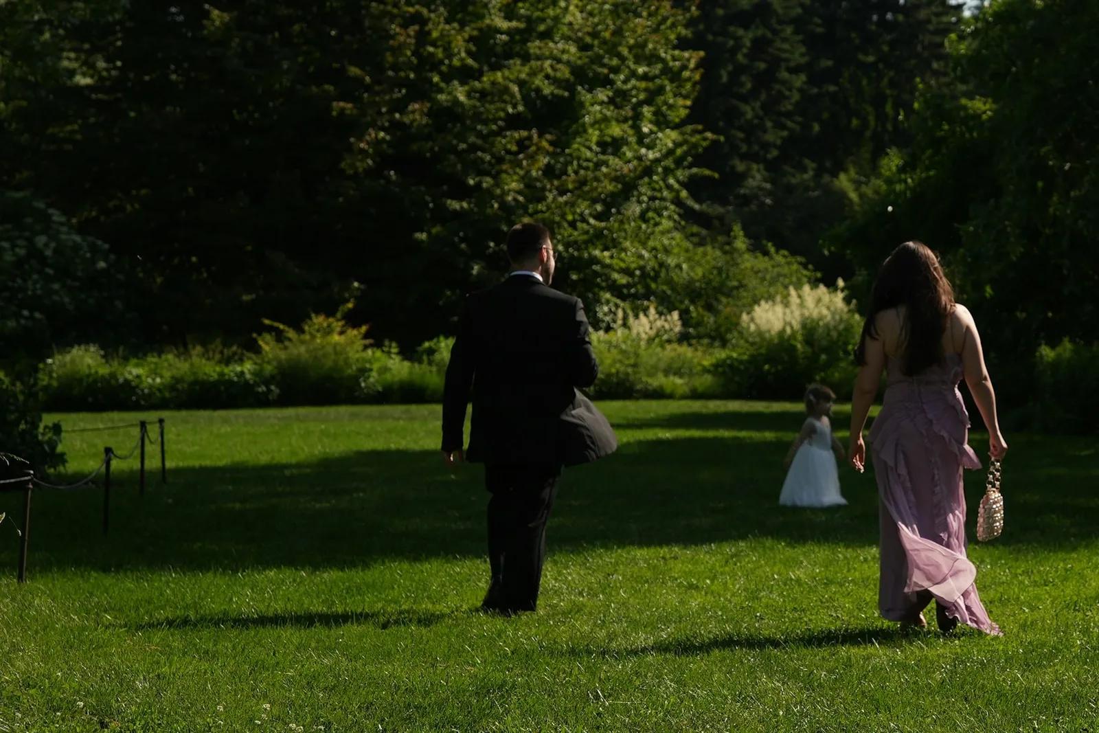 Dos invitados de boda con traje oscuro y vestido largo lila, vistos de espaldas, cruzando un césped al sol